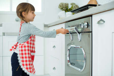 little helper. A pretty girl in a culinary apron prepares pizza, puts the dough in the oven in the kitchen.の写真素材