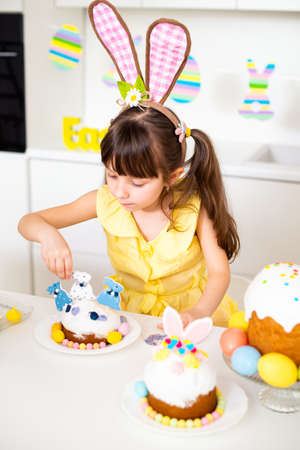 A cute little girl with bunny ears prepares an Easter cake and painted eggs. Religious holiday.