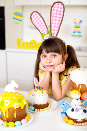 A cute little girl with bunny ears prepares an Easter cake and painted eggs. Religious holiday.