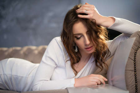Pretty girl in a white dress is lying on the sofa with a fashion magazine. high quality photoの写真素材