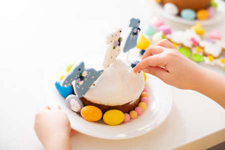 Close-up of child's hands decorating Easter cake with festive decor, sugar rabbit figurines and...