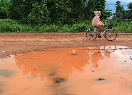 Young woman riding bicycle in orange raincoat along orange puddle on coutryside ground road on a summer day horizontal view 1の写真素材