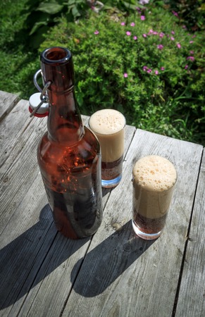brown glass bottle and two tall glasses full of frothy dark beer on rustic wooden table in summer garden, selective focusの写真素材
