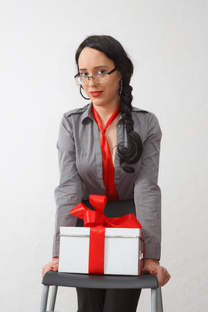 Young sexy business lady standing by chair with gift box with red ribbon on itの写真素材