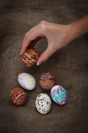 Colorful Easter eggs on burlap, female hand chose and pick one, top view close up