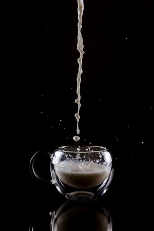 Pouring process of milk into a glass cup with reflection under glass cup, splashes and drops around glass cup against black backgroundの写真素材