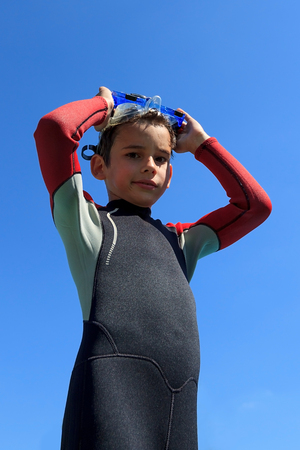 Young boy ready for diving in diving suit puts on diving mask, portrait against blue sky on sunny...