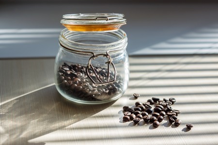Coffee seeds in transparent glass container on white table with shadows from the blinds on sunny morning, horizontal viewの写真素材