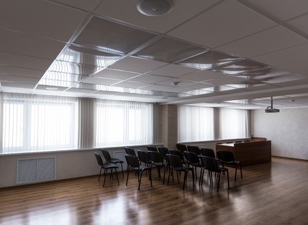 Empty sunlit modern meeting room with projector hanging on ceiling and black chairs standing in...