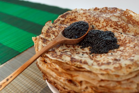 Pile of homemade pancakes with black caviar on top and wooden spoon on white plate on table covered by tablecloth, horizontal viewの写真素材