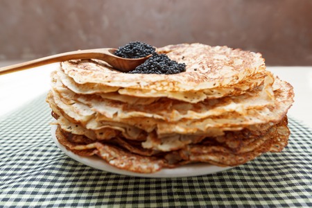 Pile of homemade pancakes with black caviar on top and wooden spoon on white plate on table covered...