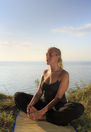 Beautiful young woman practising yoga on mat outdoors at river bank on grass, close upの写真素材