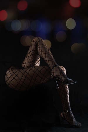 Low key photo of sexy slim beautiful legs in black net tights sitting against black background with colorful blursの写真素材