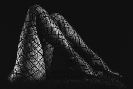 Low key greyscale photo of sexy slim beautiful legs in black net tights and high heels shoes against black background, horizontal viewの写真素材