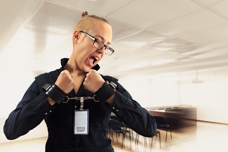 Young beautiful businesswoman screaming with anger in office, prisoner of job in cuffs, horizontal viewの写真素材