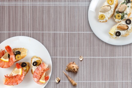 Tasty various italian sandwiches with seafood against bamboo mat background. Crostini with cheese, king shrimps, lemon, sliced olives on white plate, horizontal top viewの写真素材