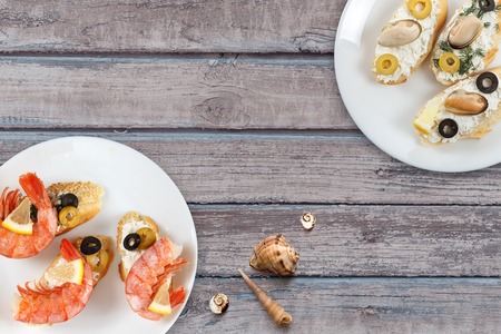 Tasty various italian sandwiches with seafood against rustic wooden background. Crostini with cheese, king shrimps, lemon, sliced olives on white plate, horizontal top viewの写真素材