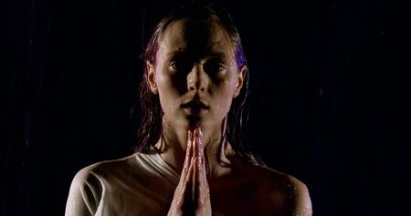 sad woman is standing with folded hands on chest under water flows in darkness, worshipping