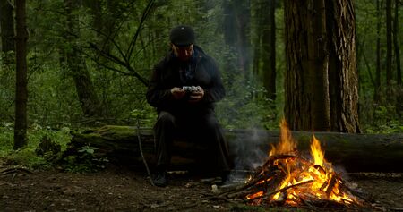 aged man sits on fallen tree trunk at bonfire with cameraの写真素材