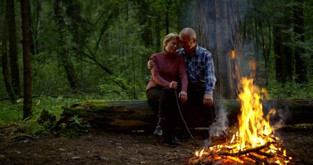 aged couple hugs and admide fire sitting on fallen treeの写真素材