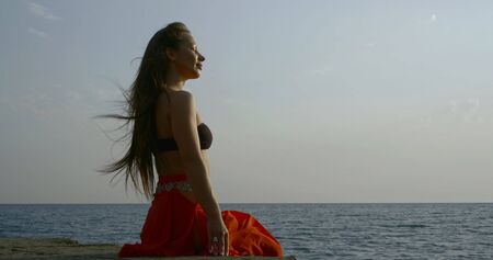 sexy woman is sitting on concrete embankment on seacoast in daytime, enjoying ocean breeze