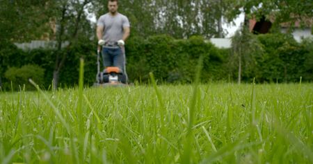 young man in t-shirt and working gloves uses modern lawn mower to cut green grass in garden low angle shot slow motionの写真素材