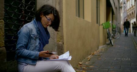 dark-haired fashionable woman in glasses middle-aged, she sits leaning against the city wall of the house, holding a mobile phone and paper, something checks and horrified.の写真素材