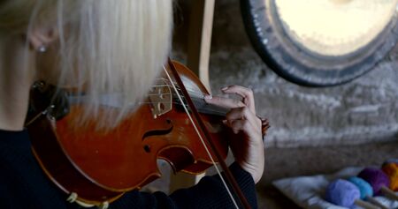 Close-up portrait of a blonde woman sitting on a stone surface and conducts sound therapy, massage sound vibrations, playing the violin, standing around her tambourines.の写真素材