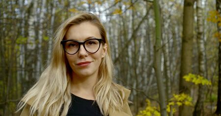 blonde woman with braces and glasses is smiling looking at camera, standing in forest in fall day