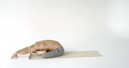 A flexible and plastic male yogi with a beard does yoga on a rug in a White Studio. He sits in the...