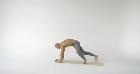 A flexible and plastic male yogi with a beard does yoga on a rug in a white Studio. He does does the...