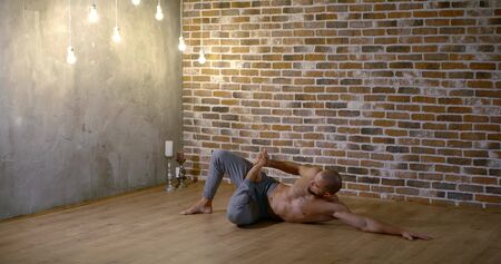 Muscular flexible bearded man doing yoga in the hall against the brick wall, he is lying on his side...