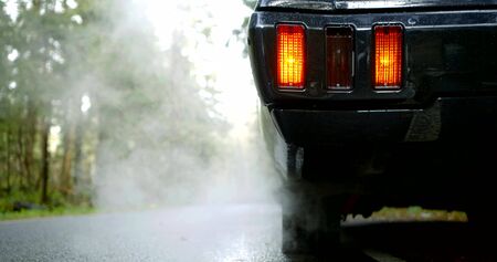 black car with lighted red tail light stands emitting white dense smoke from exhaust pipe slow...