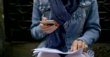 a woman with a blue scarf around her neck sits on a bench with a phone and documents. checks information on documents and phone. red nail Polish. close upの写真素材