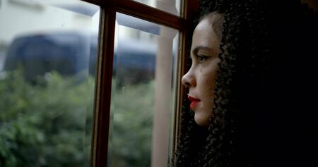 sad pensive girl with curly hair stands at the window in the roomの写真素材