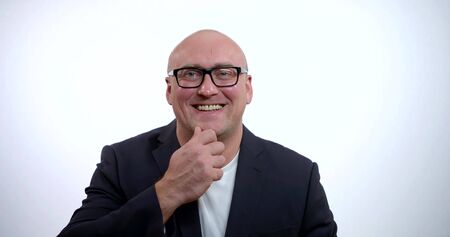 A portrait of a happy, cheerful, joyful middle-aged bald businessman in glasses and a black jacket, he is in the Studio on a white background, he smiles widely, and rubs his chin with his hand.の写真素材