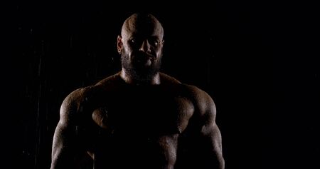Professional bodybuilder a man stands in the dark in the rain with a naked torso on his muscles...