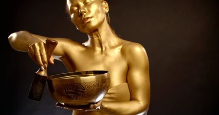 portrait of a toned girl in a top, she is in the Studio on a black background, she has a Golden pigment on her skin. She dances an Oriental dance. Creates a massage with sound vibrations using a bowl.の写真素材