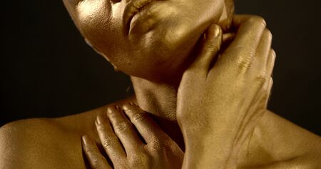 Close-up portrait of a gentle naked girl, she is in the Studio on a black background, she has a Golden pigment on her skin. She poses, makes massage movements with her hands on her shoulders.の写真素材