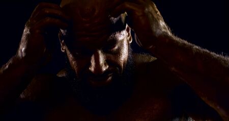portrait of a huge bald man with bronze skin on a black background. he rubs his head with both hands, scratches his beard, stares at the camera, turns in profile. dark keyの写真素材
