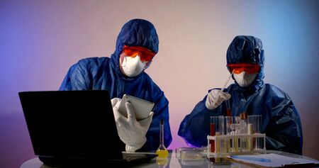 Two virologist researchers in protective clothing sit in the laboratory and study substances in test...