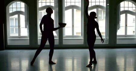 A young slender blonde girl is in a large Studio with large Windows, she is holding a bowl and a stick, creating a massage with sound vibrations, and in the background is another girl who is dancing.の写真素材