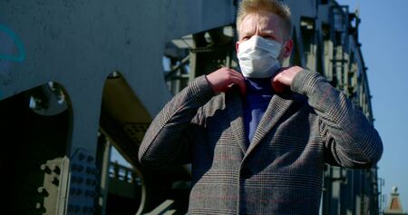 portrait of a man on a city street. he puts a disposable mask on his face, walks along the bridge...