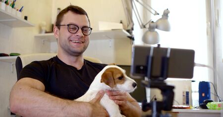 cheerful man with cute small dog is calling by video by smartphone, sitting at home
