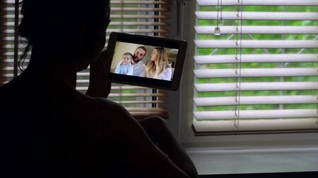 Close-up of a woman standing against the light at a window with blinds, she is talking on a video...