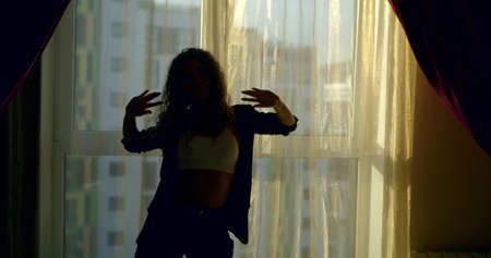 a curly-haired young beautiful girl who is standing in front of a large window, wearing a white...