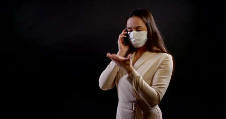 businesswoman in a protective mask stands against a black background and speaks emotionally on the phoneの写真素材