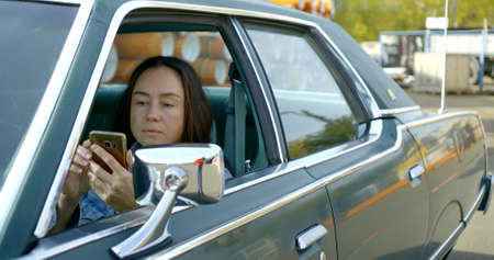 Portrait of a dark-haired woman in a denim jacket sitting behind the wheel of a classic American car...