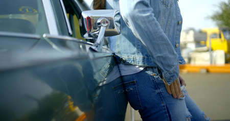 Close-up of a woman in a denim jacket and jeans leaning on a classic car, she puts her hand in her...