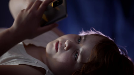 little child girl is using smartphone before asleep, lying in bedroom at night time, surfing...